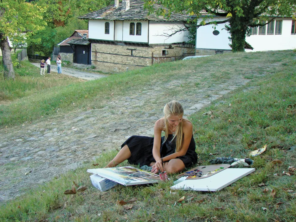 A woman painting outdoors on the grass in front of village houses