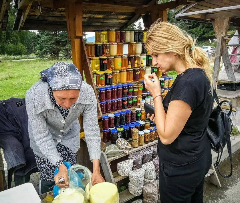 A traveller tasting at a village stall stacked with jars of honey and preserves